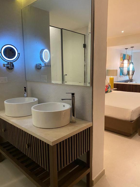 Mūsdienīgs viesnīcas numura vannas istabas interjers ar divām balta keramikas izlietnēm uz koka pamatnes un spoguli, kas apgaismots ar modernām gaismām. Fonā redzams guļamgalds ar krāsainām dekorācijām.