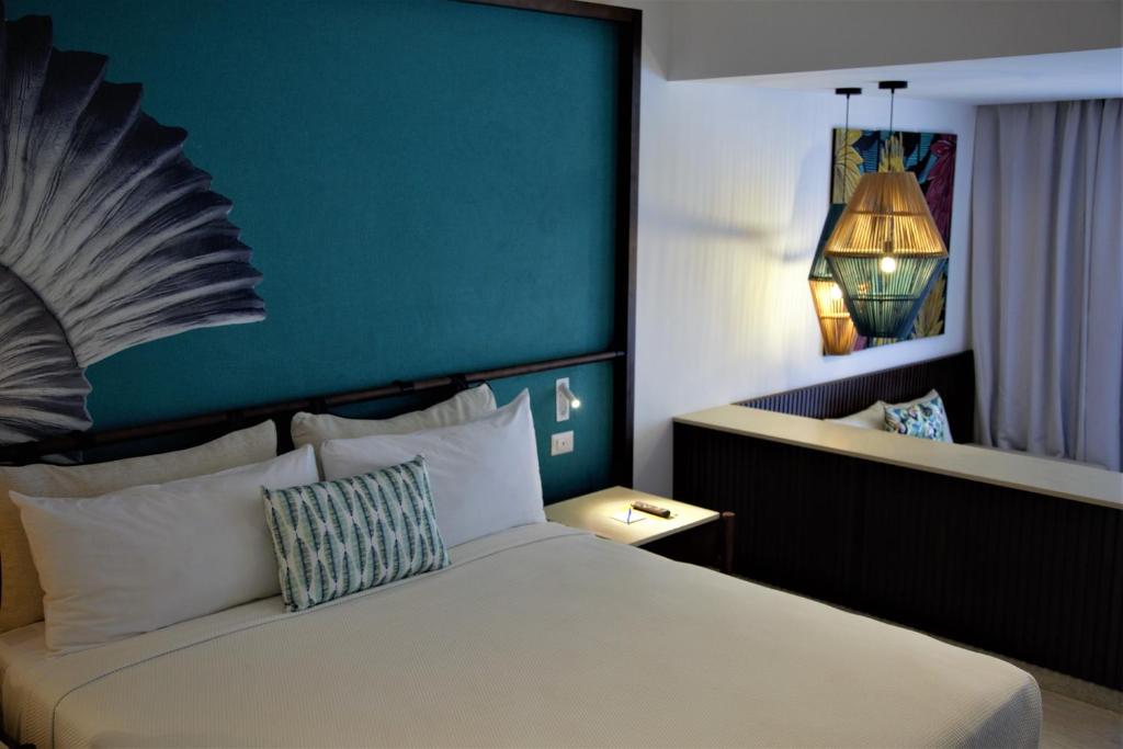 Mūsdienīgs viesnīcas numurs ar stilīgu interjeru, kas ietver zaļganu sienu un pievilcīgas gaismas lampas. Gulta ir ērti aprīkota ar spilveniem un dekoratīvu spilvenu.