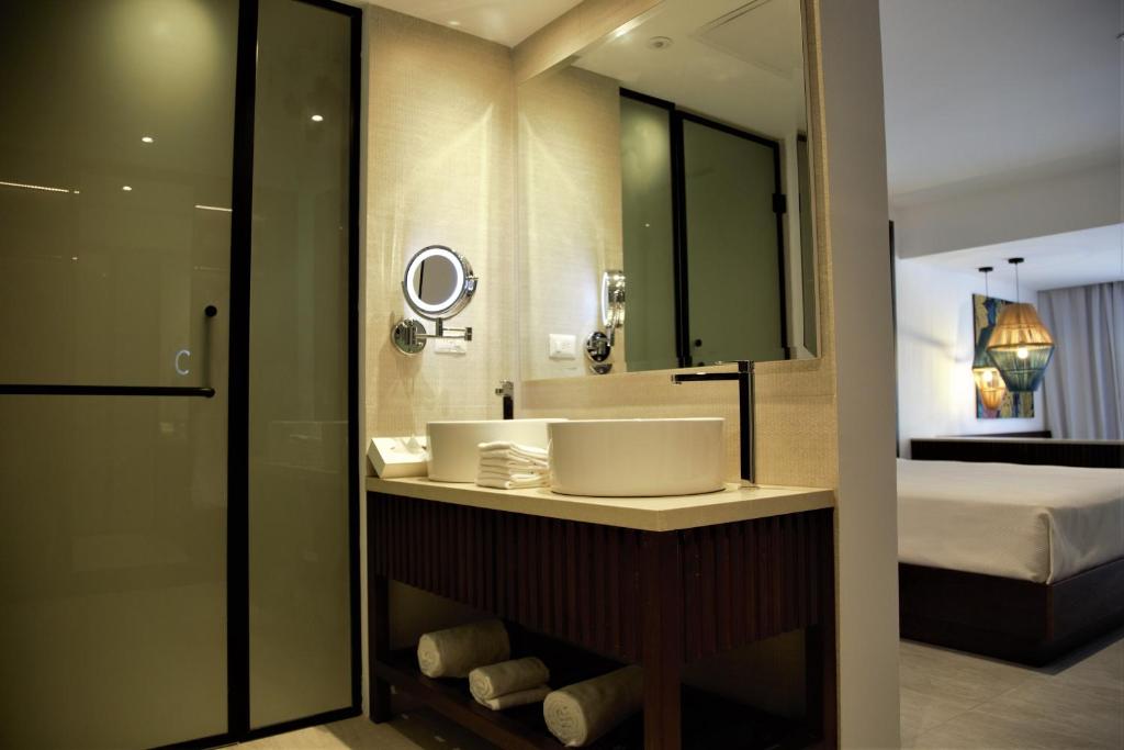 Mūsdienīgs vannas istabas interjers ar baltu izlietni un spoguli, kas atspoguļo stilīgu dizainu un ērtības. Aiz durvīm redzama telpa ar gultu un apgaismojumu.