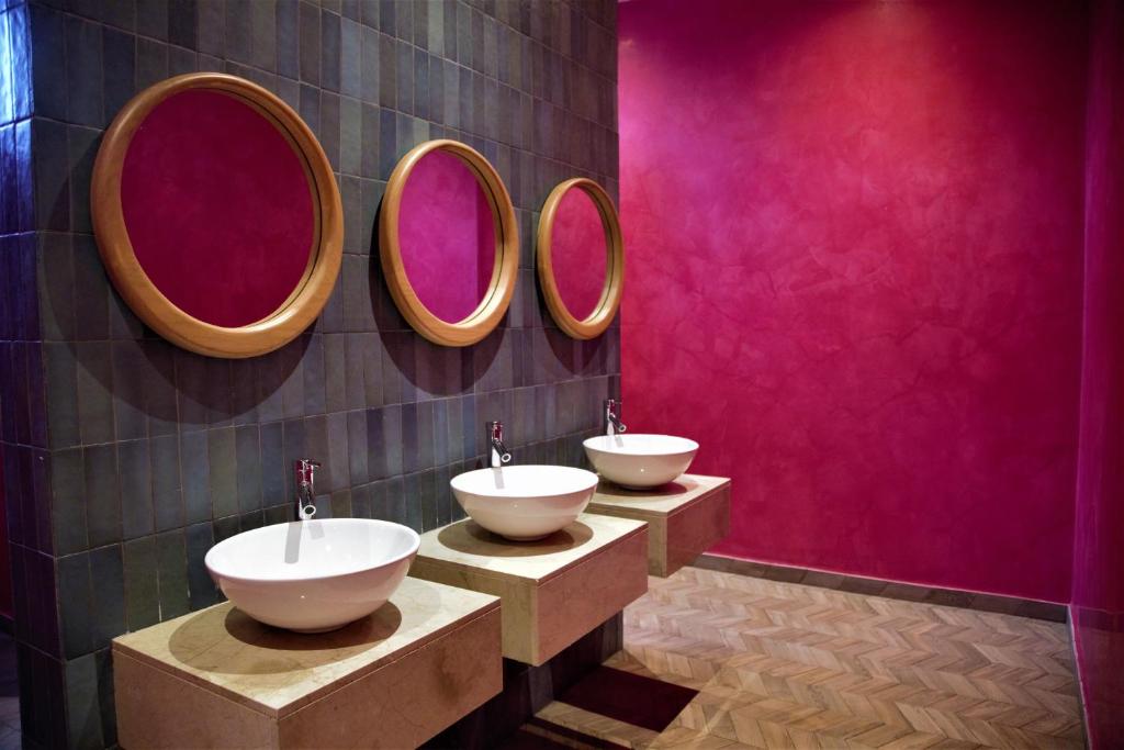 Moderns un krāsains vannas istabas interjers ar trim baltiem izlietnēm un apaļām spoguļiem, kas uzsver dzīvīgu rozā sienu.