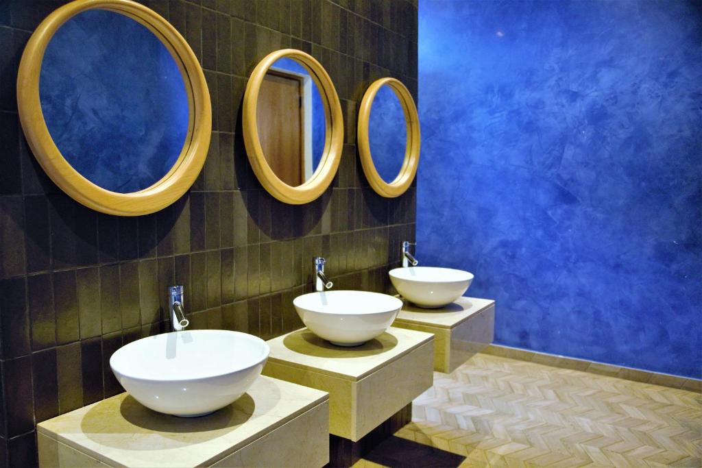 Moderns vannas istabas interjers ar trīs baltiem izlietnēm un apaļām koka spoguļiem uz tumša flīžu fona. Aiz tām ir gaiši zila siena, radot elegantu un relaksējošu atmosfēru.