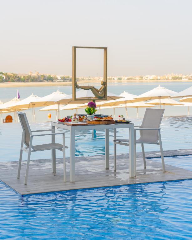 Elegantiņa galda un krēslu komplekts uz ūdens, piedāvājot brokastis ar augļiem un ziediem. Fons ar saulainu pludmali un saulessargiem.