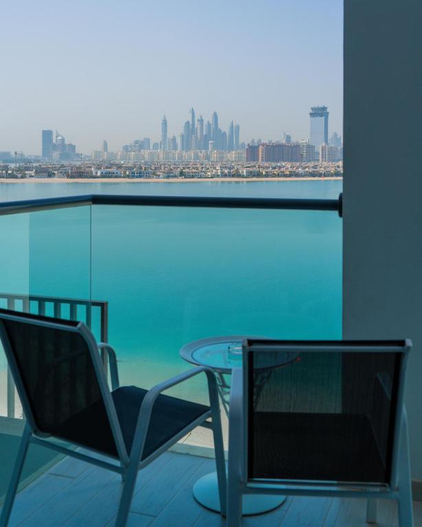 Skats no balkona uz Dubaijas pilsētas panorāmu, kur redzami moderni debesskrāpji un mierīga zila ūdens virsma. Balkonā ir divas krēsli un neliela galdiņa.