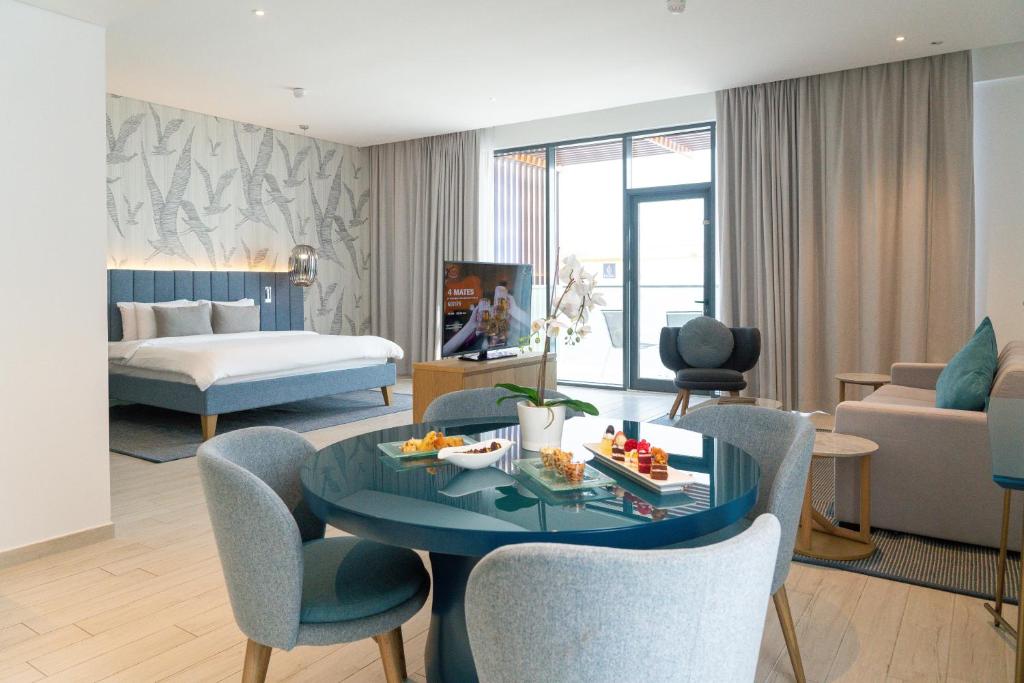 Mūsdienīgs viesnīcas numurs ar atvērtu plānojumu, kas ietver guļamistabu, dzīvojamo zonu un ēdamgaldu. Interjers ir gaišs, ar stilīgām mēbelēm un dabīgām krāsām, radot komfortablu atmosfēru.