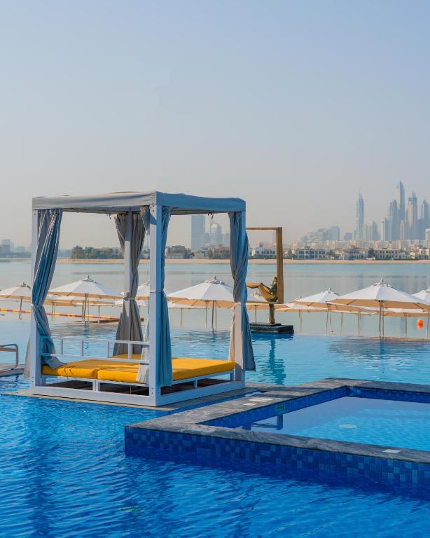 Peldbaseins ar baltu kanālu un zeltainu atpūtas gultu, fonā redzama pilsētas ainava ar augstceltnēm un sauļošanās vietām.