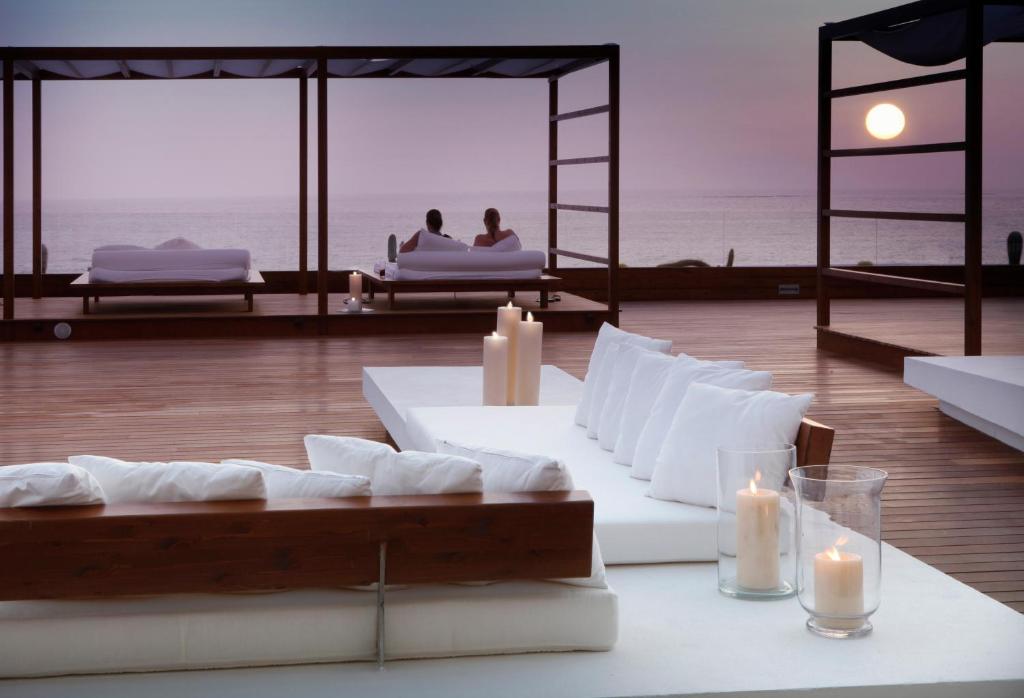 Romantisks skats uz jūru ar mīkstām baltām spilveniem un sveču gaismām, radot mierīgu atmosfēru saulrieta laikā.