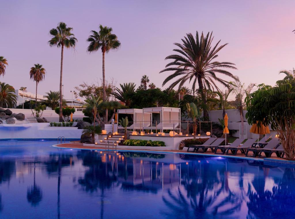 Luksusa baseins ar palmu kokiem un mīkstām sauļošanās vietām, kas atrodas blakus modernai viesnīcai, radot relaksējošu atmosfēru saulrieta gaismā.