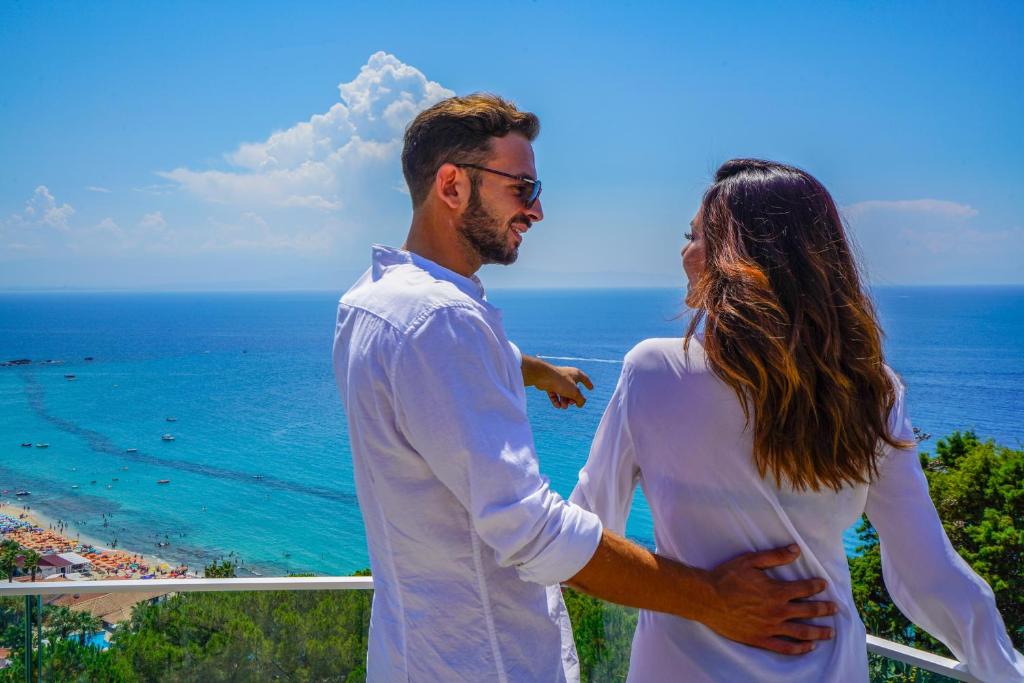 Jauns pāris, apģērbts baltās drēbēs, skatās uz jūru no terases ar skatu uz pludmali un laivām. Gaisa zils un saulains, radot romantisku atmosfēru.