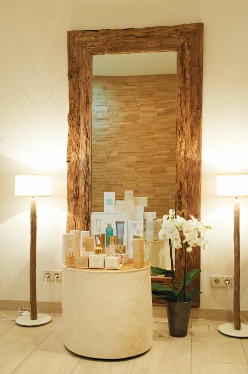 Elegants interjers ar spoguli, kas ietverts masīvā koka rāmī, un stilīgiem apgaismojuma ķermeņiem. Uz apaļas galda izvietoti dažādi kosmētikas produkti, papildināti ar baltu orhideju podiņā.