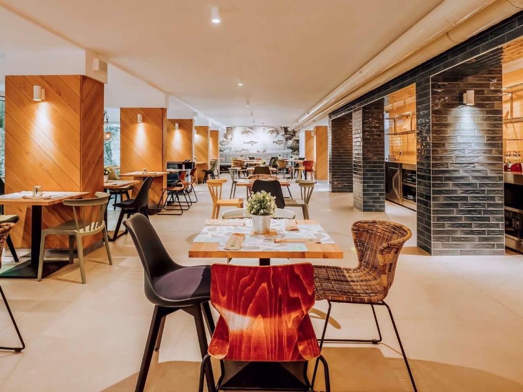 Mūsdienīgs restorāna interjers ar koka un metāla elementiem, dažādu krāsu krēsliem un galdiem, kas rada siltu un aicinošu atmosfēru. Apgaismojums akcentē telpas modernās līnijas un ērtību.