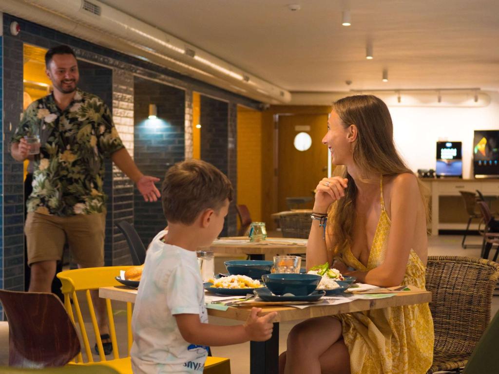 Bērns un sieviete sēž pie galda, baudot maltīti, kamēr vīrietis ar dzērienu rokā tuvojas. Gaisotne ir draudzīga un relaksēta, ar modernu restorāna iekārtojumu.