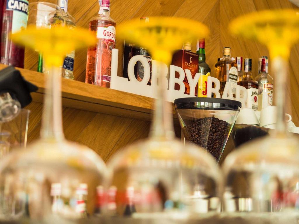 Bāra skats ar dzērienu pudelēm un izsmalcinātiem glāzēm priekšplānā, uz koka plaukta redzams bāra nosaukums "LOBBY BAR".