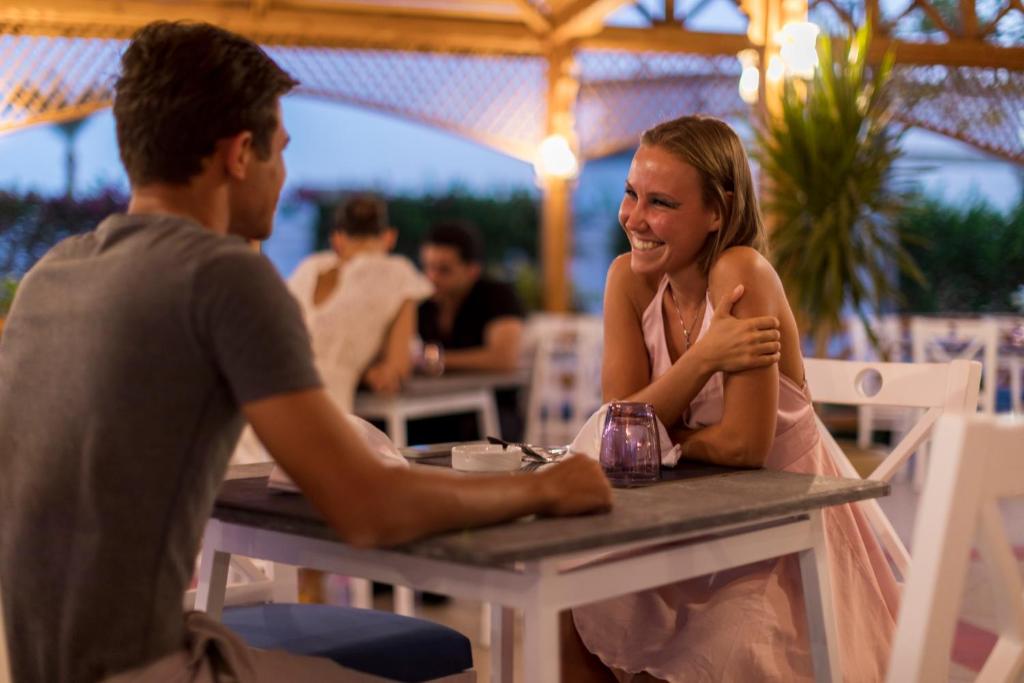 Divi cilvēki sēž pie galda restorānā, priecīgi sarunājoties un smejoties. Apkārt redzami citi apmeklētāji un gaisma no lampām rada patīkamu atmosfēru.