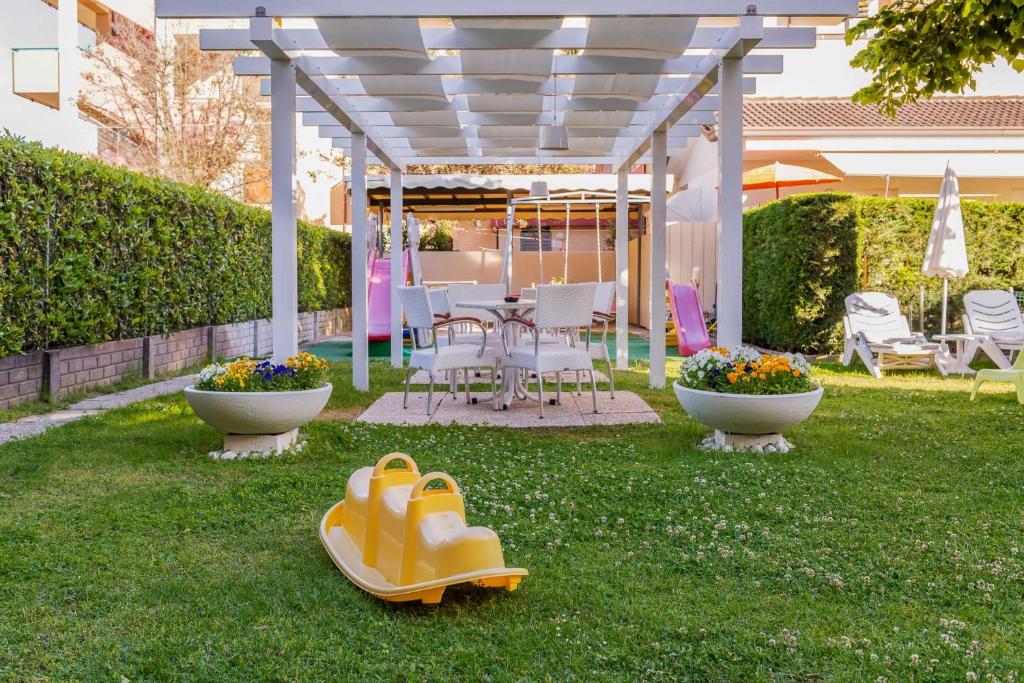 Dārza zona ar balto pergolu, sēdvietām un dzeltenu slīdkalniņu, ko ieskauj ziedoši puķu podi. Ideāla vieta relaksācijai un spēlēm.