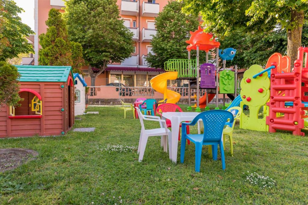 Krāšņs bērnu rotaļu laukums ar plastmasas slide, kāpņu konstrukcijām un rotaļu mājiņu, apkārt zaļojoša zāle un krāsainas krēsli.