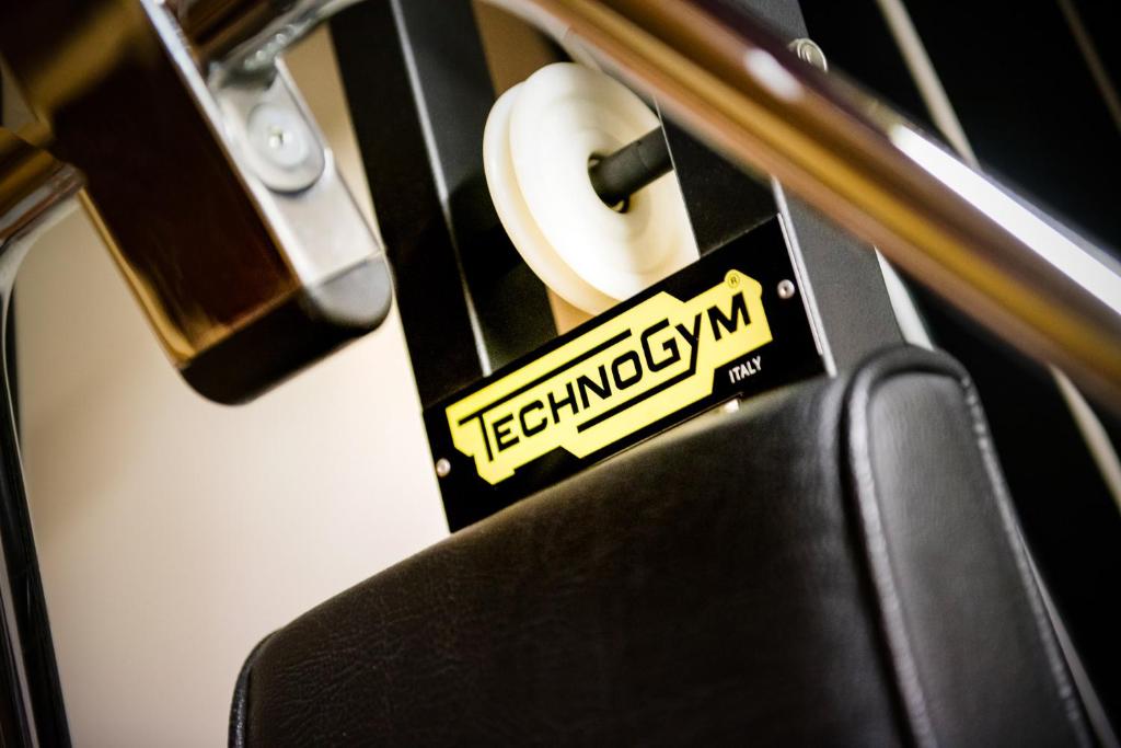 Tehnoloģiskās sporta iekārtas detaļa ar "Technogym" zīmolu un melnu sēdekli, kas norāda uz augstas kvalitātes fitnesa aprīkojumu.