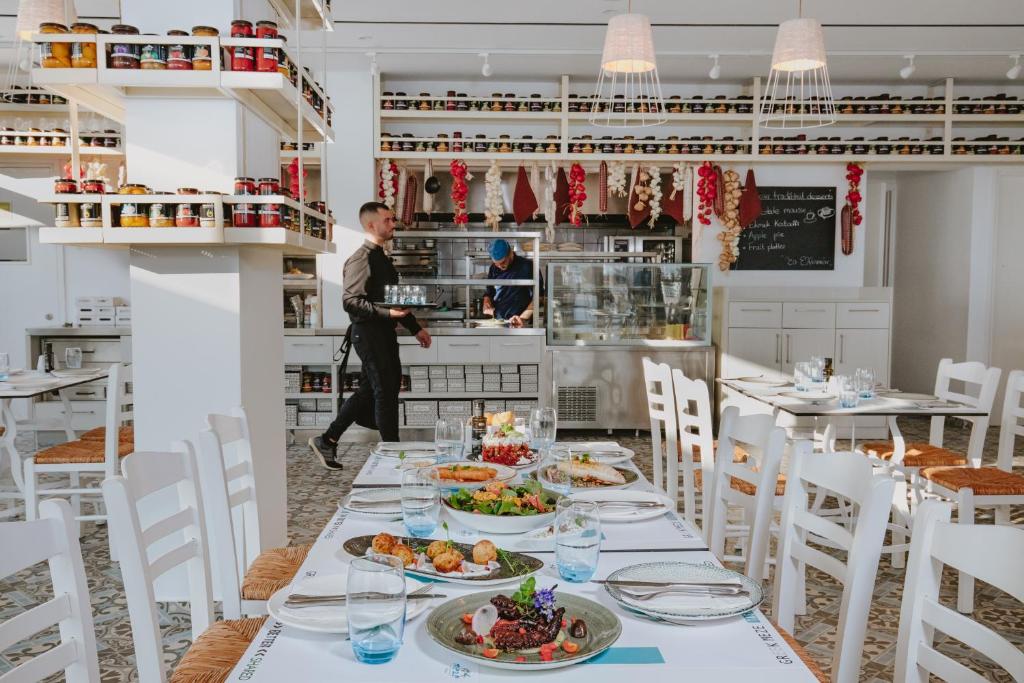 Mūsdienīga restorāna iekšskats ar elegantām galdiem, kas klāti ar dažādiem ēdieniem un dzērieniem. Aiz galdiem redzams pavārs, kas strādā virtuvē, un plaukti ar konservētiem produktiem un garšvielām.