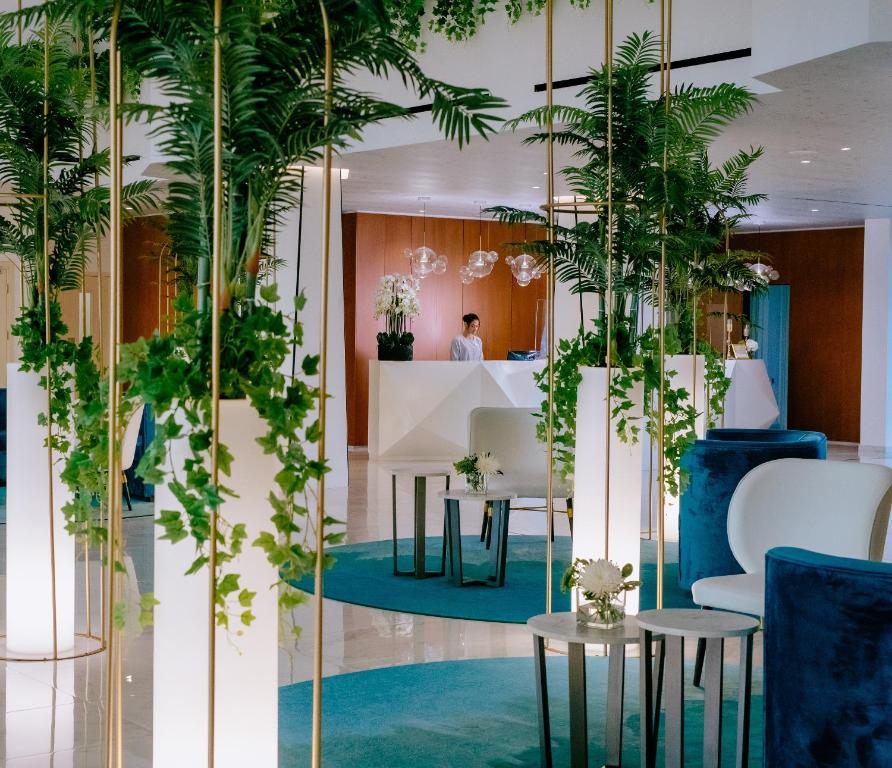 Mūsdienīgs viesnīcas interjers ar zālēm un elegantiem mēbeļu elementiem, kas rada relaksējošu atmosfēru. Skatāms reģistratūras galds un dekoratīvas augu kompozīcijas.