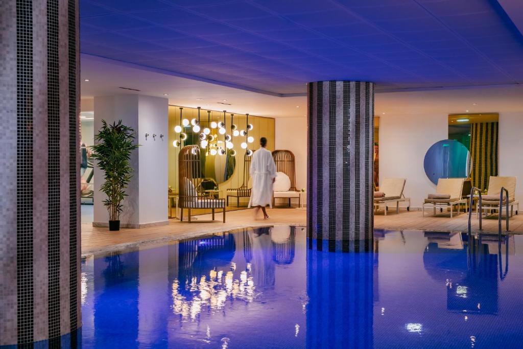 Mūsdienīgs spa iekārtojums ar zilu ūdeni un relaksējošu atmosfēru, kurā redzama persona baltā drānā, dodoties uz atpūtas zonu.