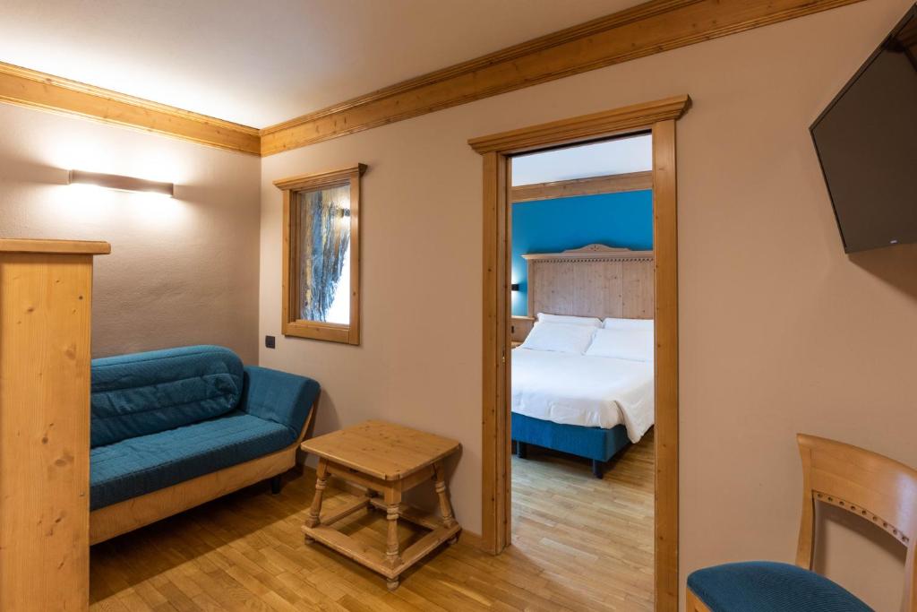 Mūsdienīgs viesnīcas numurs ar koka apdari, iekārtots ar zilu dīvānu un koka galdiņu, redzama guļamistaba ar divguļamo gultu.