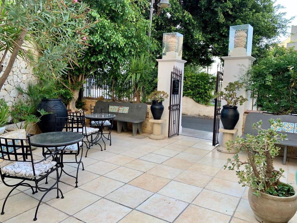 Skaista dārza zona ar galdiem un krēsliem, kas izveidota relaksējošai atmosfērai. Vārti ar skulptūrām un zaļām augiem, kas veido viesmīlīgu ieeju.