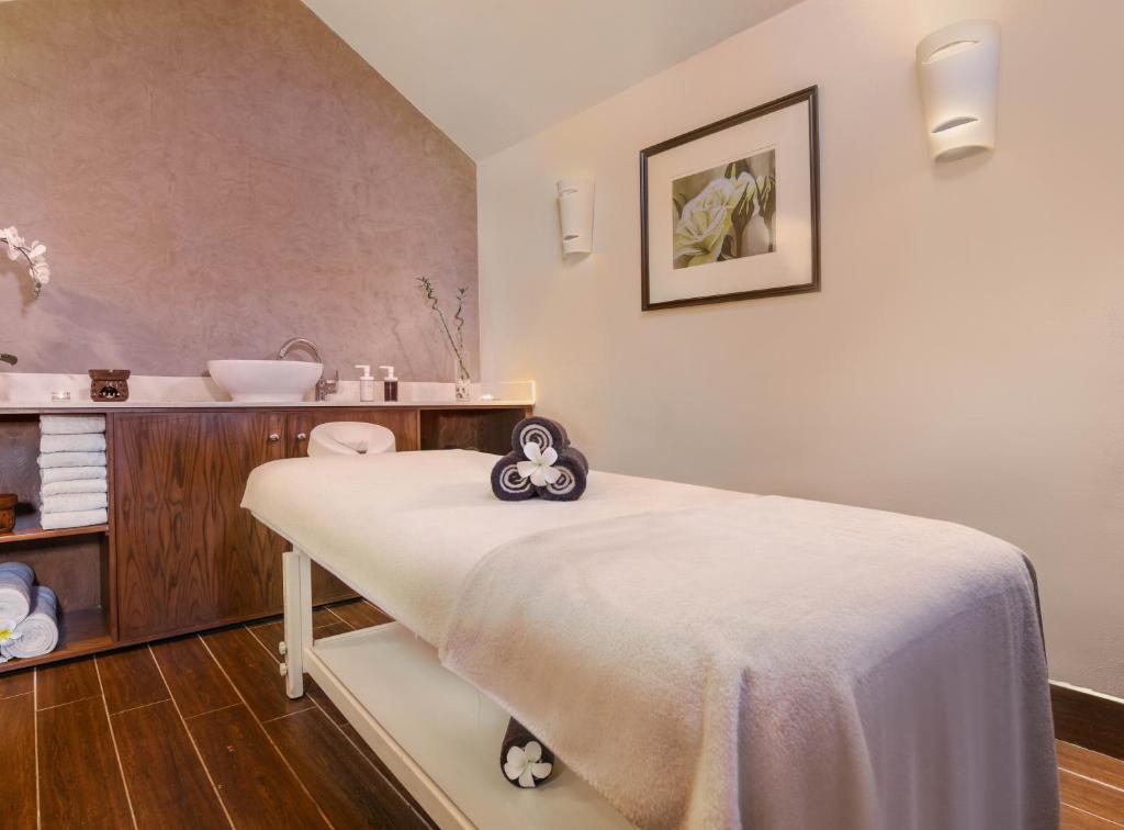 Mūsdienīgs spa telpa ar masāžas gultu, stilīgiem dvieļiem un relaksējošu atmosfēru. Interjerā ir koka mēbeles un dekoratīvs zieds, kas veicina mieru un atpūtu.
