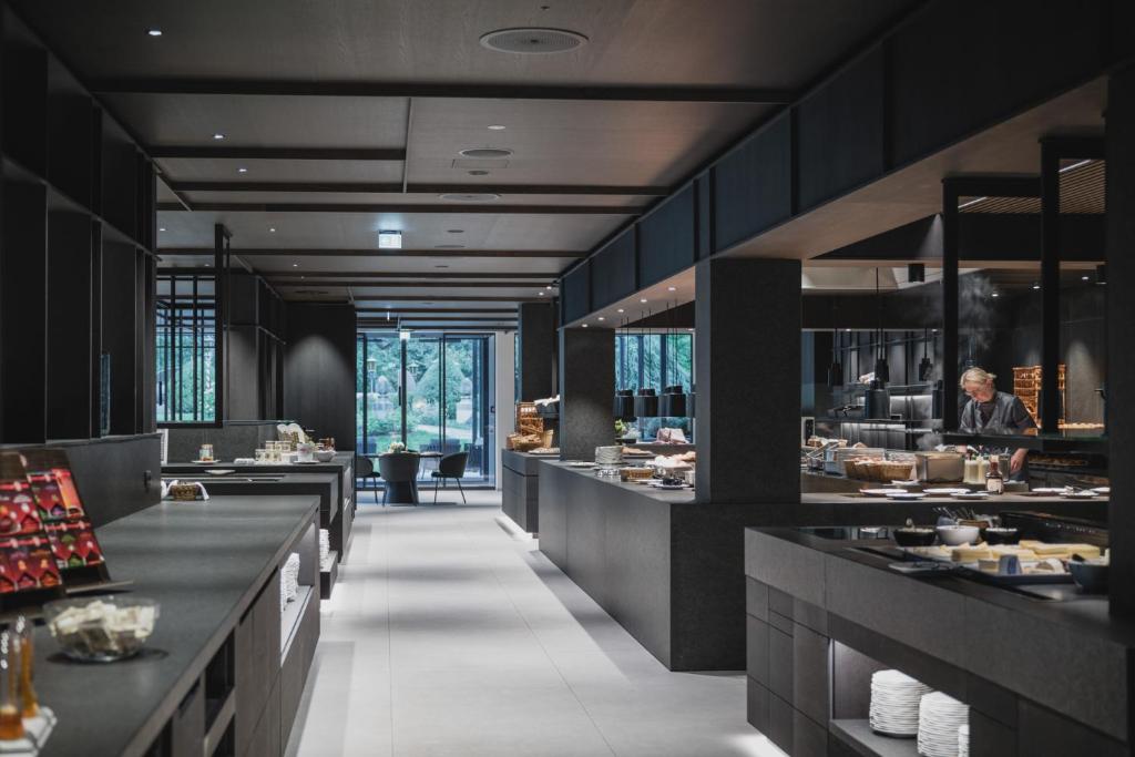 Moderna un eleganta restorāna iekārtojums ar gaišām telpām un melniem elementiem, kur šefpavārs gatavo ēdienus. Redzamas dažādas uzkodas un dzērieni uz galdiem.