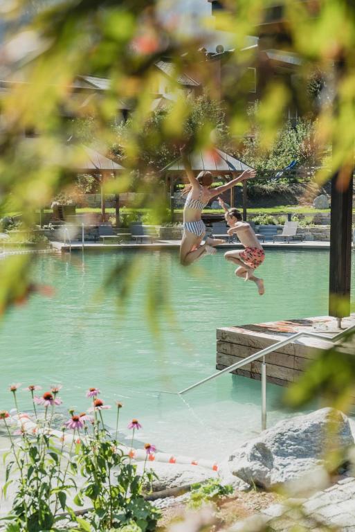 Divi bērni lēkā ūdenī no mola, priecīgi izbaudot vasaras brīvdienas. Apkārt ir zaļš dīķis un ziedošas puķes, kas piešķir ainavai dzīvīgumu.