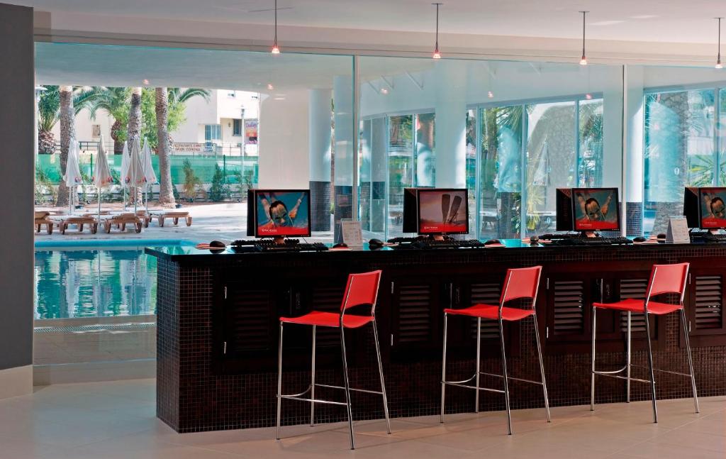 Mūsdienīgs bārs ar sarkaniem krēsliem un datoriem, kas atrodas blakus baseinam, ar palmām un sauļošanās krēsliem redzamā fonā. Ideāla vieta relaksācijai un izklaidei.
