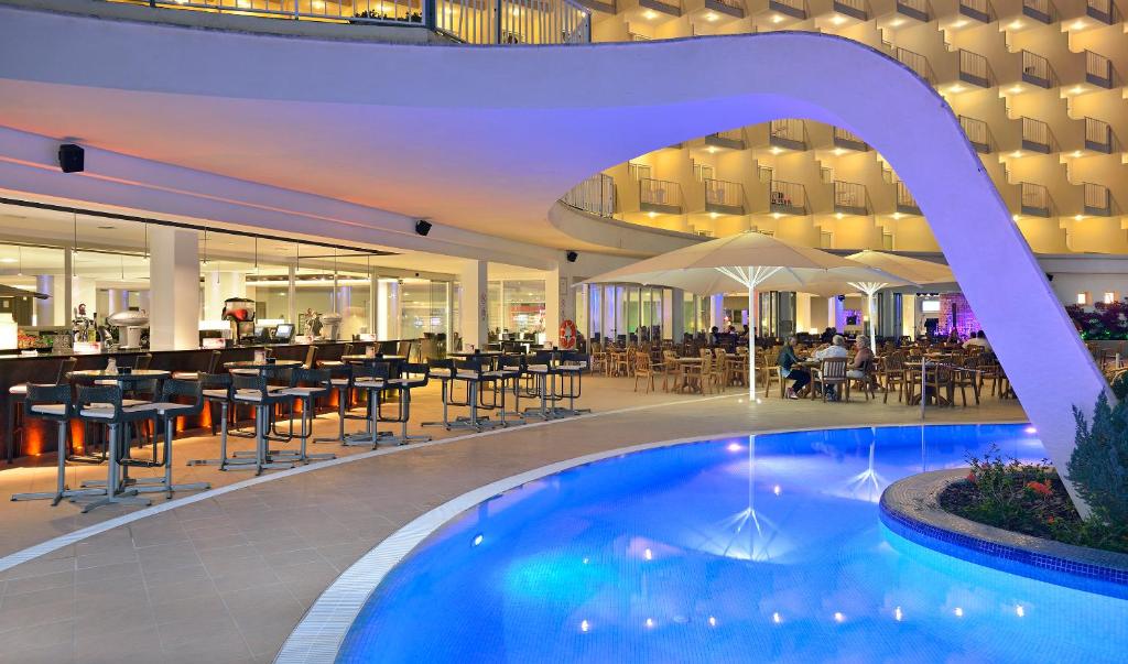 Mūsdienīgs viesnīcas bārs ar elegantu peldbaseinu, apgaismotu ar zilu gaismu, un redzamu restorāna zonu. Šī ainava rada relaksējošu atmosfēru atpūtai un izklaidei.