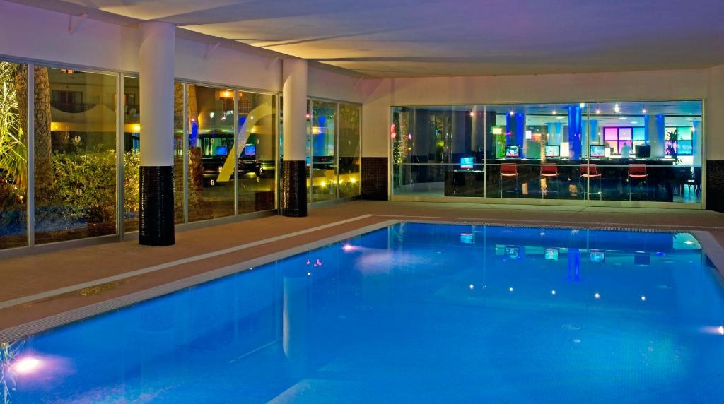 Mūsdienīgs iekštelpu baseins ar dzidru zilu ūdeni un spilgtu apgaismojumu, kas rada relaksējošu atmosfēru. Aiz stikla sienām redzama mūsdienīga telpa ar datoriem un krēsliem, kas piešķir modernu pieskaņu.