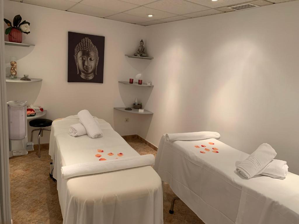 Mierīgas masāžas telpas interjers ar diviem masāžas galdiem, dekorētiem ar rožu ziedlapiņām, un budas attēlu uz sienas, radot relaksējošu atmosfēru.