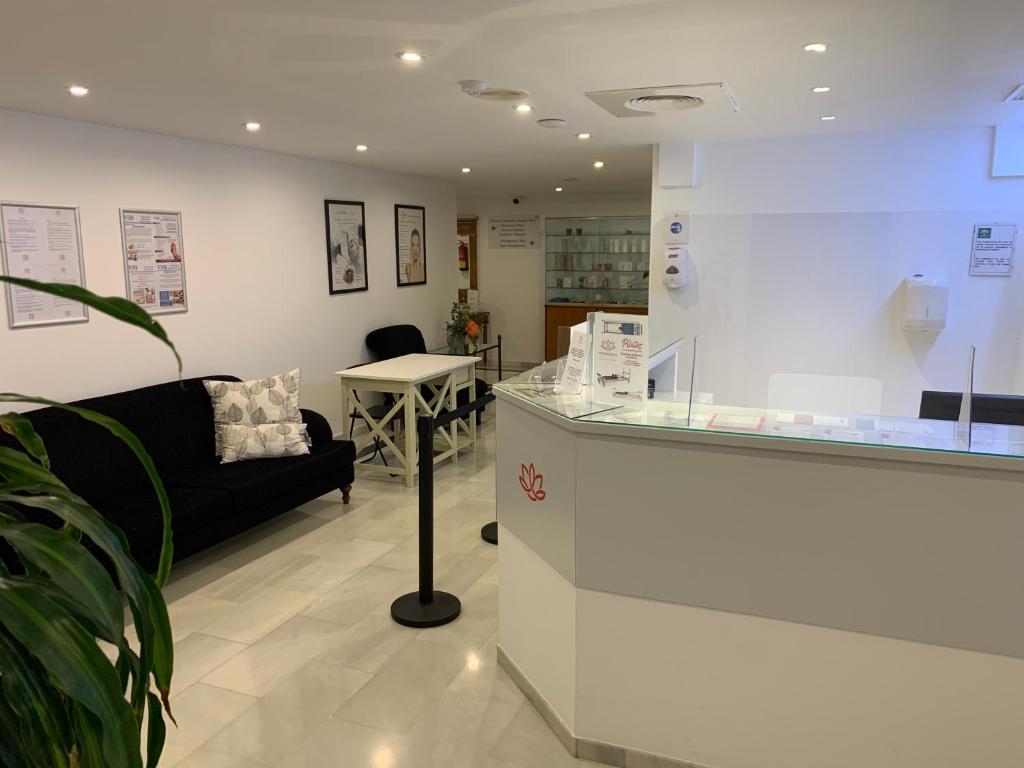 Mūsdienīgs un gaišs reģistratūras interjers, kurā redzama melna dīvāna zona ar spilveniem un baltu galdu, kā arī skaidri redzamais reģistratūras galds ar informatīviem materiāliem.