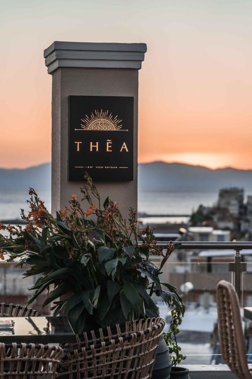 Elegants restorāna "THĒA" zīme ar saulrietu fonā, kas piešķir patīkamu atmosfēru. Apskatāmas zaļas augu kompozīcijas un moderna terase.