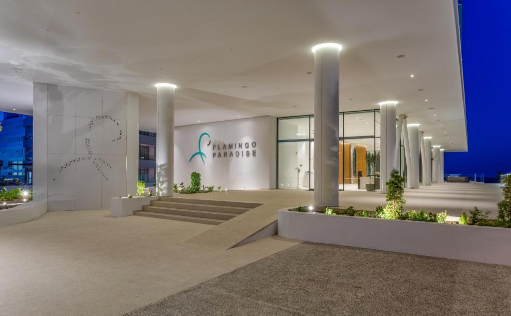 Mūsdienīgs viesnīcas ieeja ar stilīgu arhitektūru un apgaismojumu, kas uzsver "Flamingo Paradise" zīmolu. Apkārt ēkai ir zaļumi un kāpnes, kas ved uz iekšpusi.