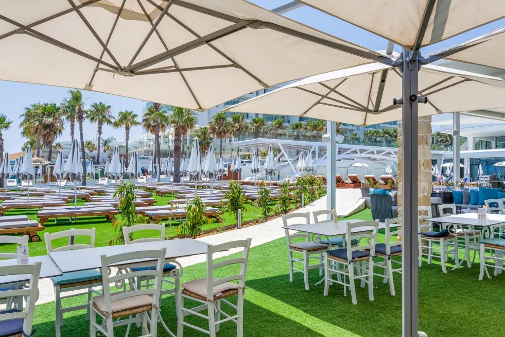 Āra restorāna terase ar baltiem galdamiem un krēsliem, ko ieskauj zaļa zāle un palmām, ar skatu uz sauļošanās gultām un pludmales saulessargiem. Ideāla vieta atpūtai un ēšanai.