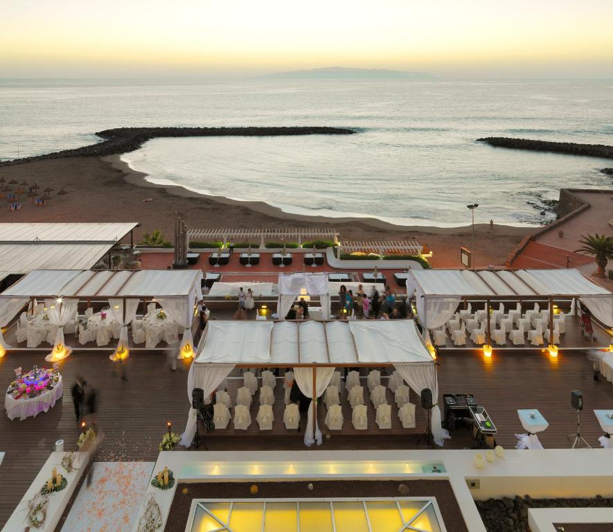 Kāzu svinības pie jūras ar elegantām teltīm un krēslu rindām, kas gaida viesus, saulrieta gaismā. Apskatāma pludmale un mierīga jūra, radot romantisku atmosfēru.