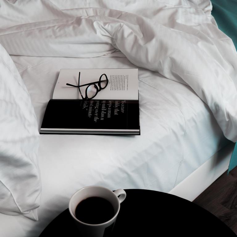 Gulta ar baltu gultasveļu, uz kuras atrodas grāmata un brilles, blakus ir kafijas krūze uz melna galda.