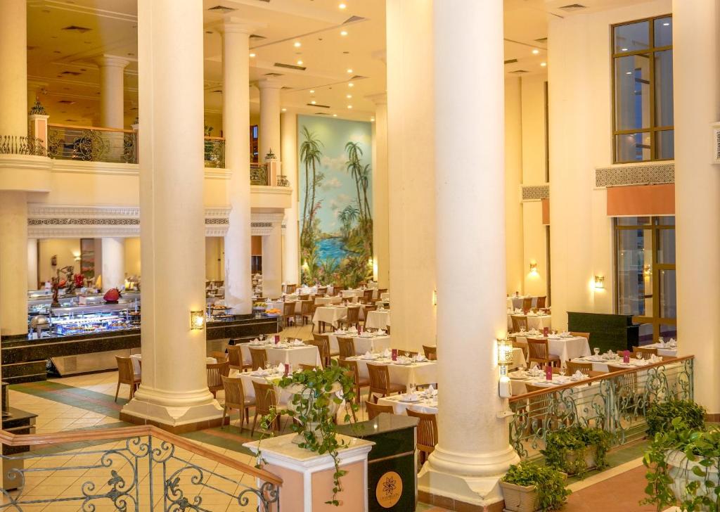 Lielas un elegantas restorāna telpas ar gaismas stariem, baltiem galdautiem un zaļiem augiem, kas rada patīkamu atmosfēru. Fons rotā krāsains attēls ar palmām pie jūras.