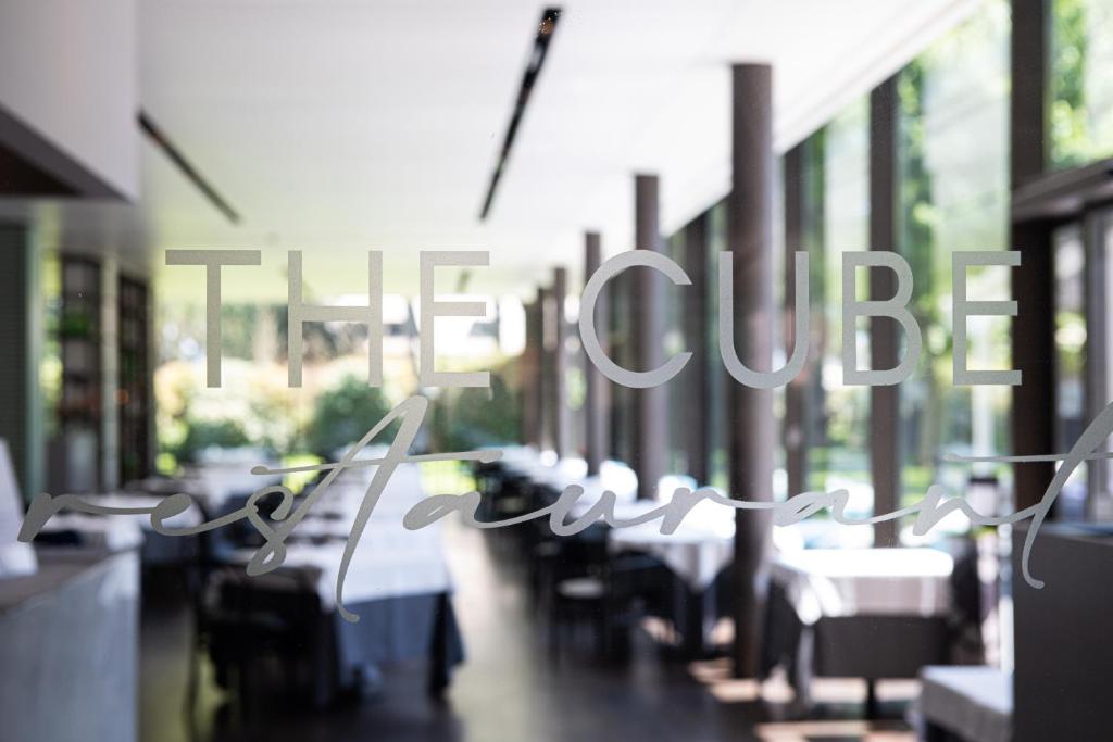 Restorāna "The Cube" nosaukums redzams caur stiklu, rādot elegantu interjeru ar galdiem un logiem, kas ved uz dārzu.
