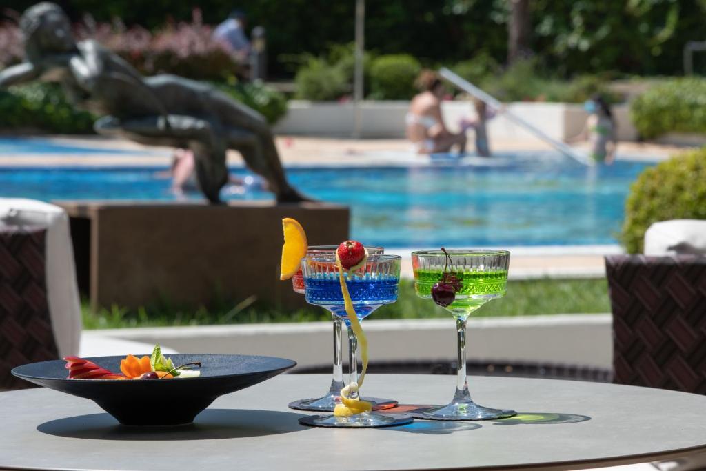 Krāšņi kokteiļi un svaigi augļi uz galda pie peldbaseina, kas izveido relaksējošu atmosfēru. Fons ietver skulptūru un cilvēkus, kas izbauda vasaras priekus.