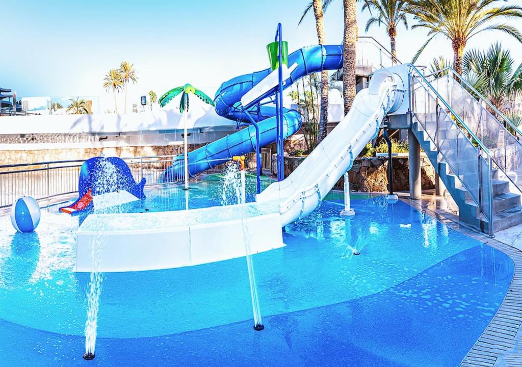Bērnu ūdens parks ar diviem ūdens slaidiem, zilu baseinu un rotaļlietu delfīnu. Apkārt aug palmām, radot tropisku atmosfēru.