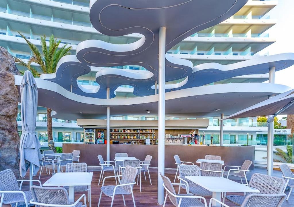 Mūsdienīgs bārs ar viļņveida jumtu un ērtām sēdvietām, kas atrodas pie baseina. Apkārt ir palmām un modernām ēkām, radot relaksējošu atmosfēru.