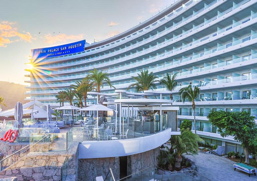 Mūsdienīgs viesnīcas komplekss ar baltiem arhitektūras elementiem un palmām, kas rada tropisku atmosfēru. Saules gaisma izgaismo ēku, uzsverot tās eleganto dizainu.