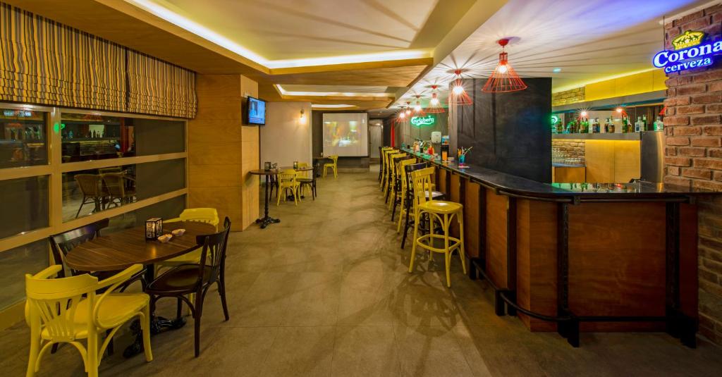 Mūsdienīgs bārs ar dzeltenām krēsliem un koka galdiem, apgaismots ar krāsainām lampām, radot patīkamu atmosfēru. Bāra plaukti ir piepildīti ar dzērieniem, un redzama televizora ekrāns.