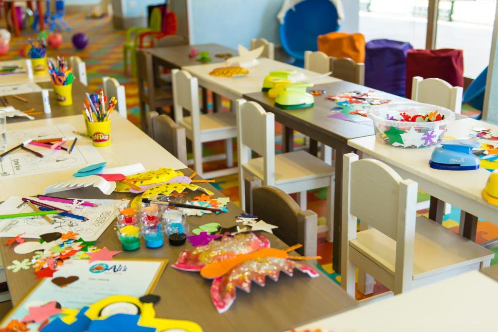 Radošas aktivitātes telpa ar krāsainām galdiem un krēsliem, kur bērni var zīmēt un veidot dažādus mākslas darbus. Uz galdiem redzama liela radošo materiālu izvēle, tostarp krāsainas papīra izgriezumi un zīmēšanas piederumi.