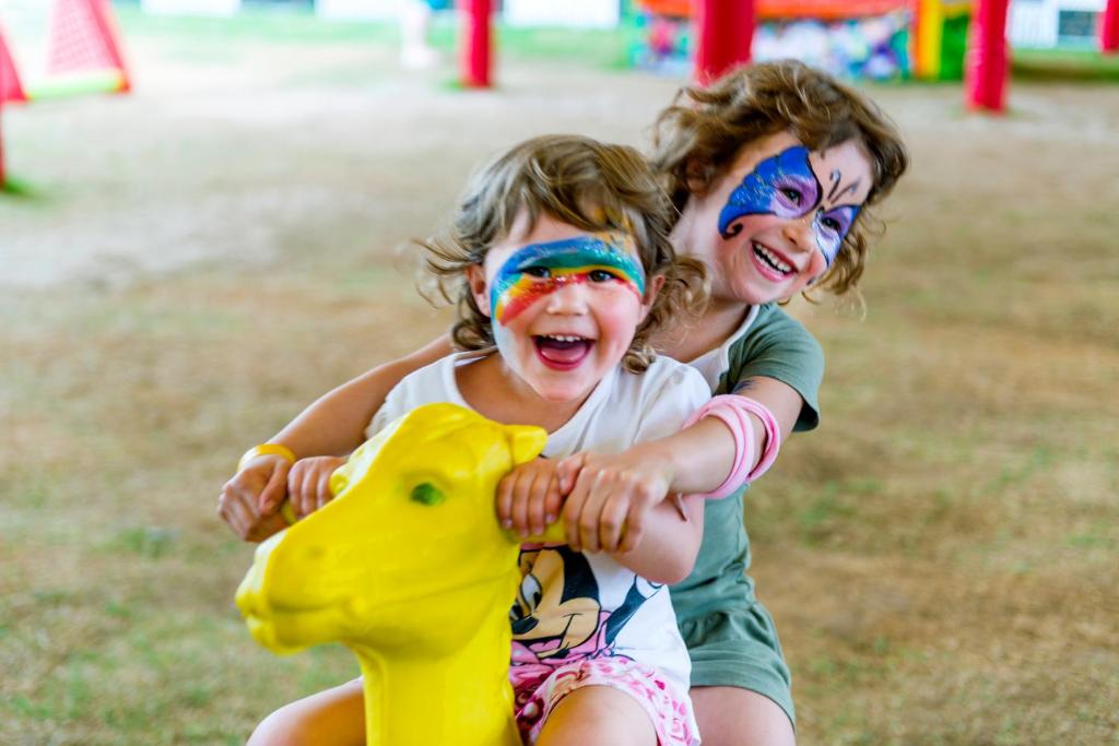 Divas priecīgas meitenes ar krāsainu sejas gleznojumu, kas sēž uz dzeltena rotaļu dzīvnieka, izsaka prieku un jautrību spēļu laukumā.