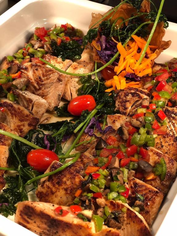 Svaiga cepta zivs ar dārzeņu garnējumu, tostarp tomātiem, papriku un lapu kāpostiem, pasniegta baltā traukā.