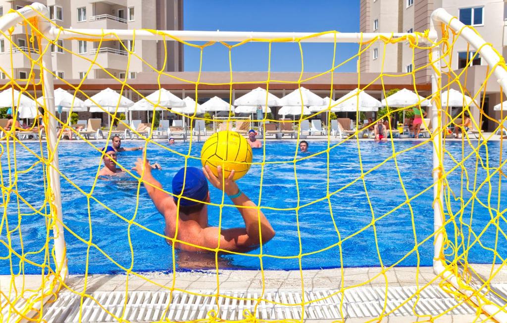 Ūdenspolo spēle baseinā, kur spēlētājs gatavojas mest bumbu pret vārtiem, kamēr citi dalībnieki ir ūdenī. Fons ir saulaina diena un atpūtas zona ar sauļošanās krēsliem un saules nojumes.