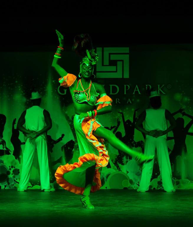 Dejotāja ar krāsainu tērpu un rotājumiem izpilda energisku deju uz skatuves, kas apgaismota ar zaļu gaismu. Fona attēlā redzami dejotāji baltos tērpos.